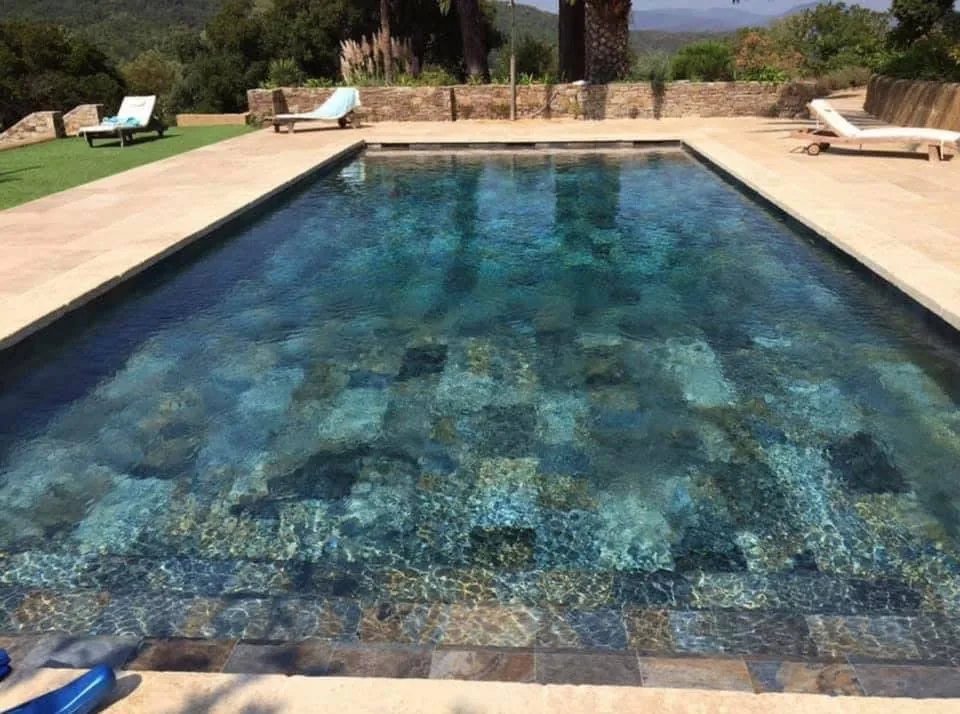 Piscine en eau carrelée en grès effet pierre aux tons bleus et ocre, entourée d'une plage en pierre claire